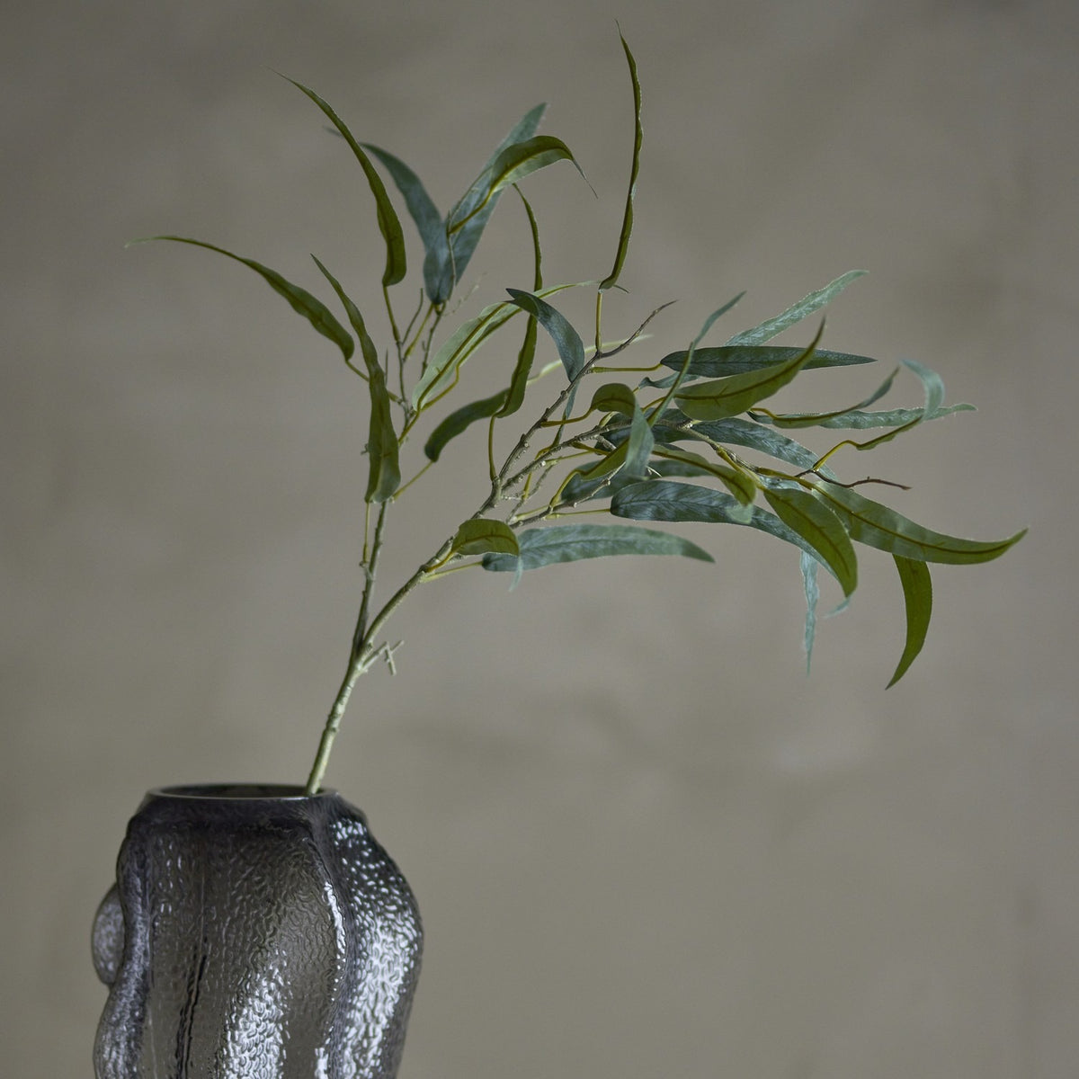 Eucalyptus Artificial Stem 85x30cm in green, a faux branch with long narrow leaves set in a textured grey glass vase against a plain grey background.