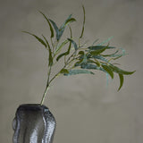 Eucalyptus Artificial Stem 85x30cm in green, a faux branch with long narrow leaves set in a textured grey glass vase against a plain grey background.