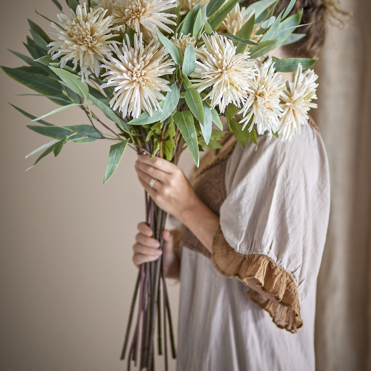 Person holding a bundled bouquet of Chrysanthemum Artificial Stem - Cream flowers with green foliage, cream petals, and long stems in a neutral indoor setting.