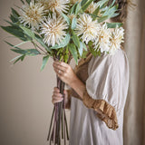 Person holding a bundled bouquet of Chrysanthemum Artificial Stem - Cream flowers with green foliage, cream petals, and long stems in a neutral indoor setting.