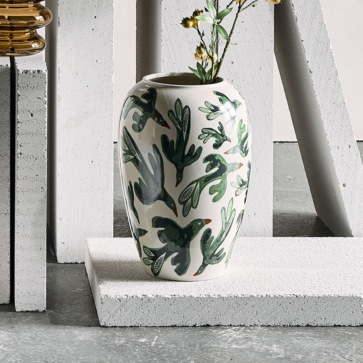 Birdliva Stoneware Vase 33cm in white with green bird and leaf motifs, rounded body and short rim, shown with foliage on a concrete plinth against a white backdrop.