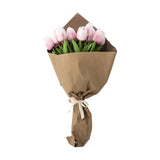 Artificial pink tulip bouquet with closed, cupped blooms and green stems wrapped in brown paper, tied with a cream bow, on a plain white background.
