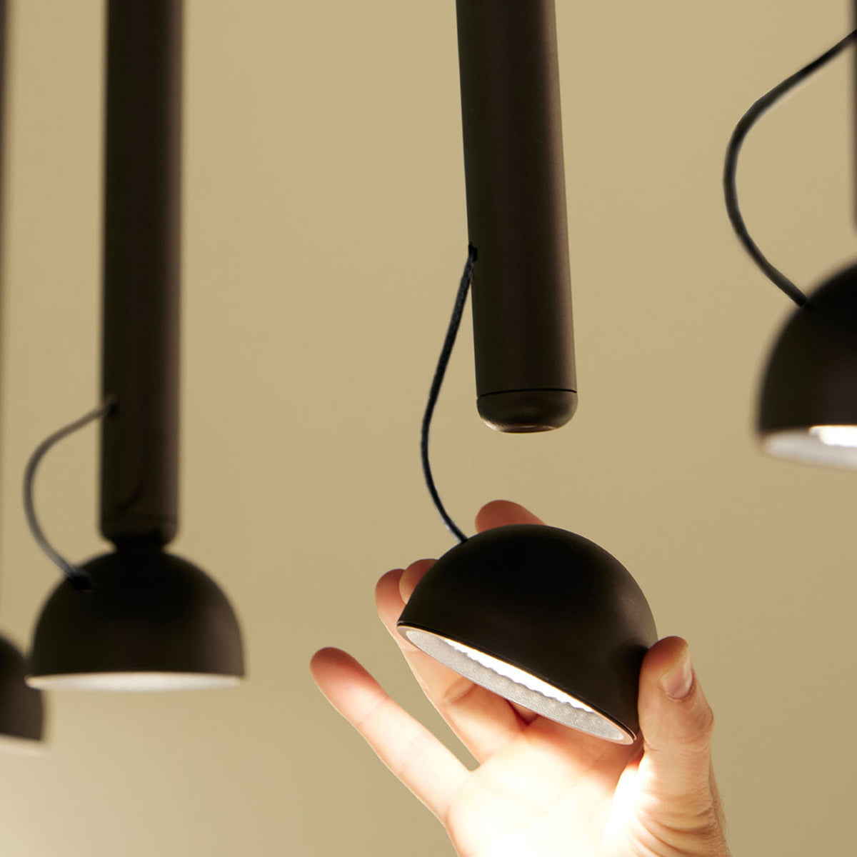 Berry S Aluminium LED pendant light in matte black, hand holding the small domed head wired to its slim tube, with similar pendants visible against a warm beige wall.