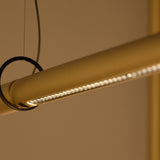 Close-up of the Solid SH Sens 220 cylinder LED pendant light, showing a slim brass-toned tube with a linear warm-white LED strip along one edge and black cable suspension.
