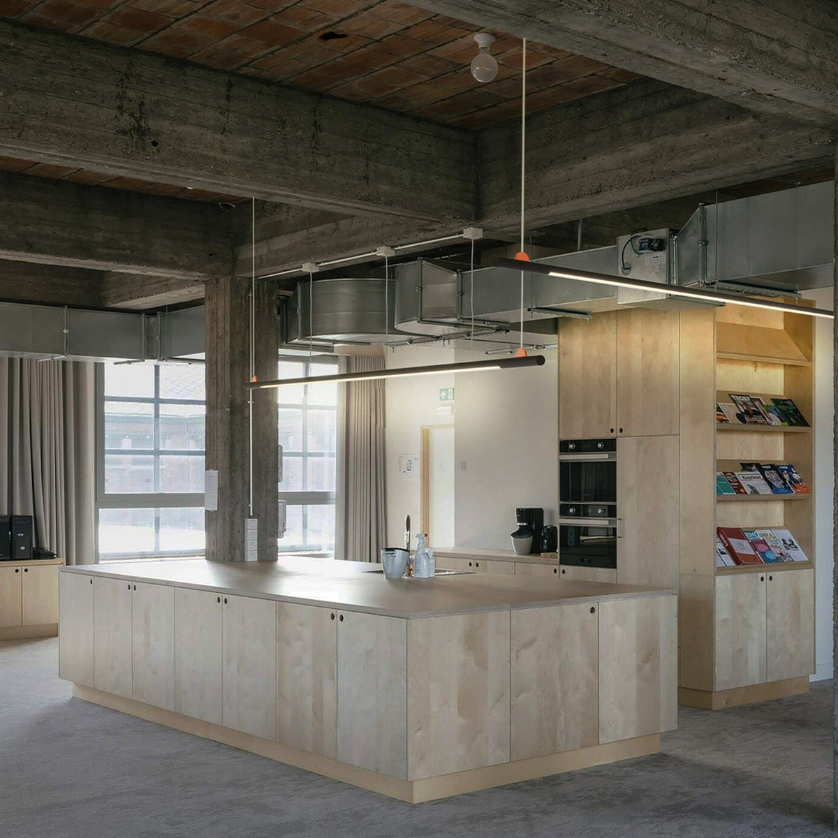 BIMU S100 Ele linear LED pendant light in black, slim bar suspended on cables above a kitchen island in an industrial-style interior with concrete ceiling.