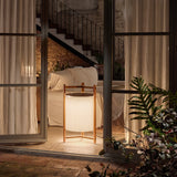 Kando P/76/R rechargeable LED floor lamp with cream pleated ribbon shade and three-legged teak wood frame, glowing in a living room doorway at night.