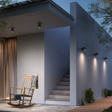 Three Nut A/01 stainless steel outdoor wall lights with bell-shaped black heads mounted on a grey exterior wall beside steps, each casting warm white downward light.