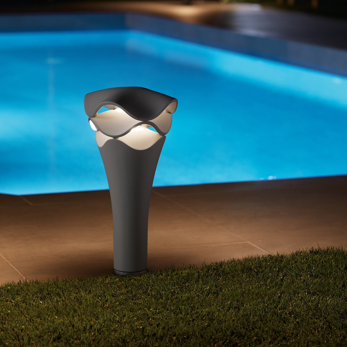 Cornet B/52 cone LED bollard light in stone grey, flared aluminium body with a wavy cap over stacked white tiers, lit warm white on grass beside a poolside patio at night.
