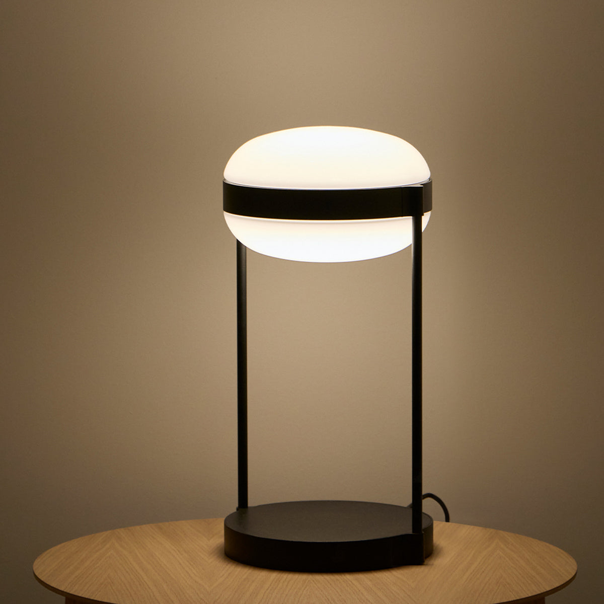 Beaded T Glass LED Table Lamp with a rounded opal white shade held by a dark band between two slim black rods on a round black base, glowing warm white on a wooden table.