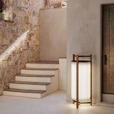 Kando P/111 Ribbon LED floor lamp with a cream pleated cylindrical shade, dark top disc, and open teak wood frame, shown by a stone-walled staircase.
