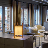 Kando M/40 Ribbon LED table lamp with pleated white shade in an open wood-toned three-legged frame, lit on a sideboard in a living room setting.