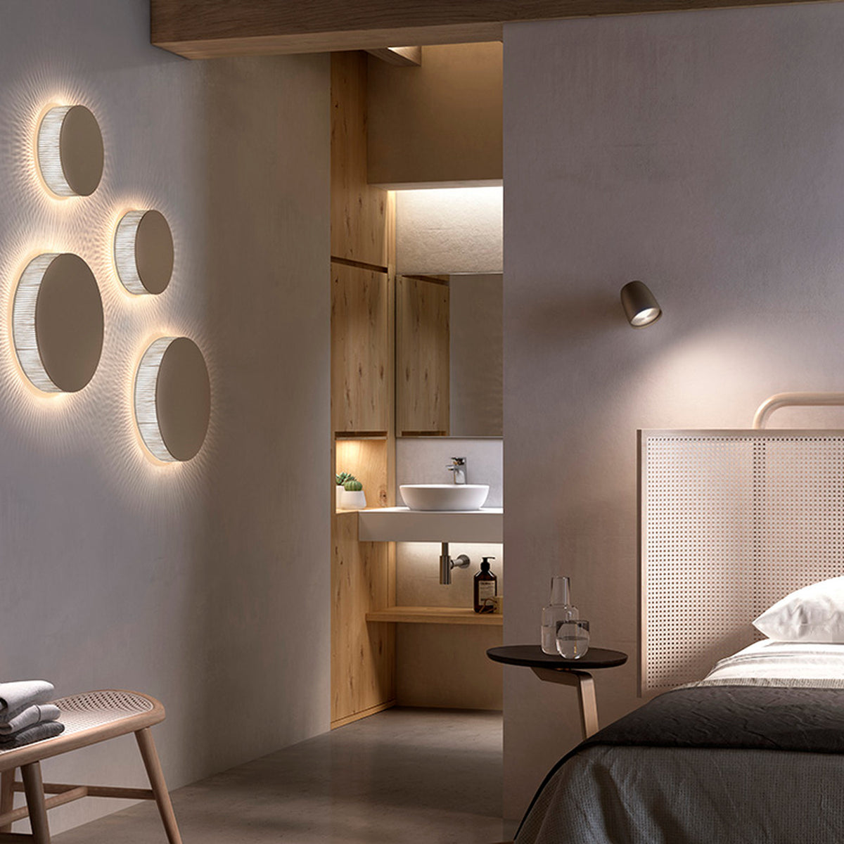Helios A/01 round LED wall lights in Oak Brown & Beige fabric, flat brown discs with glowing cotton thread edge, shown grouped on a wall above a bedroom with a bed, nightstand and adjoining bathroom.