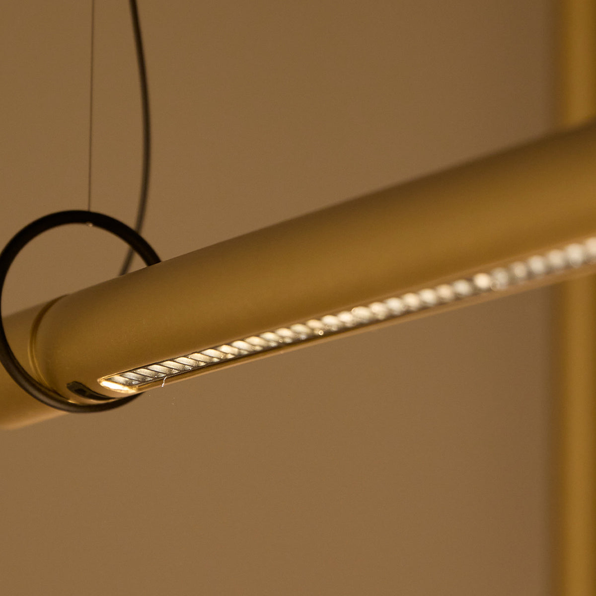 Close-up of the Solid SH Sens cylinder pendant light in brass, showing the slim tube with a single-line LED strip along its underside, suspended on fine black wires.