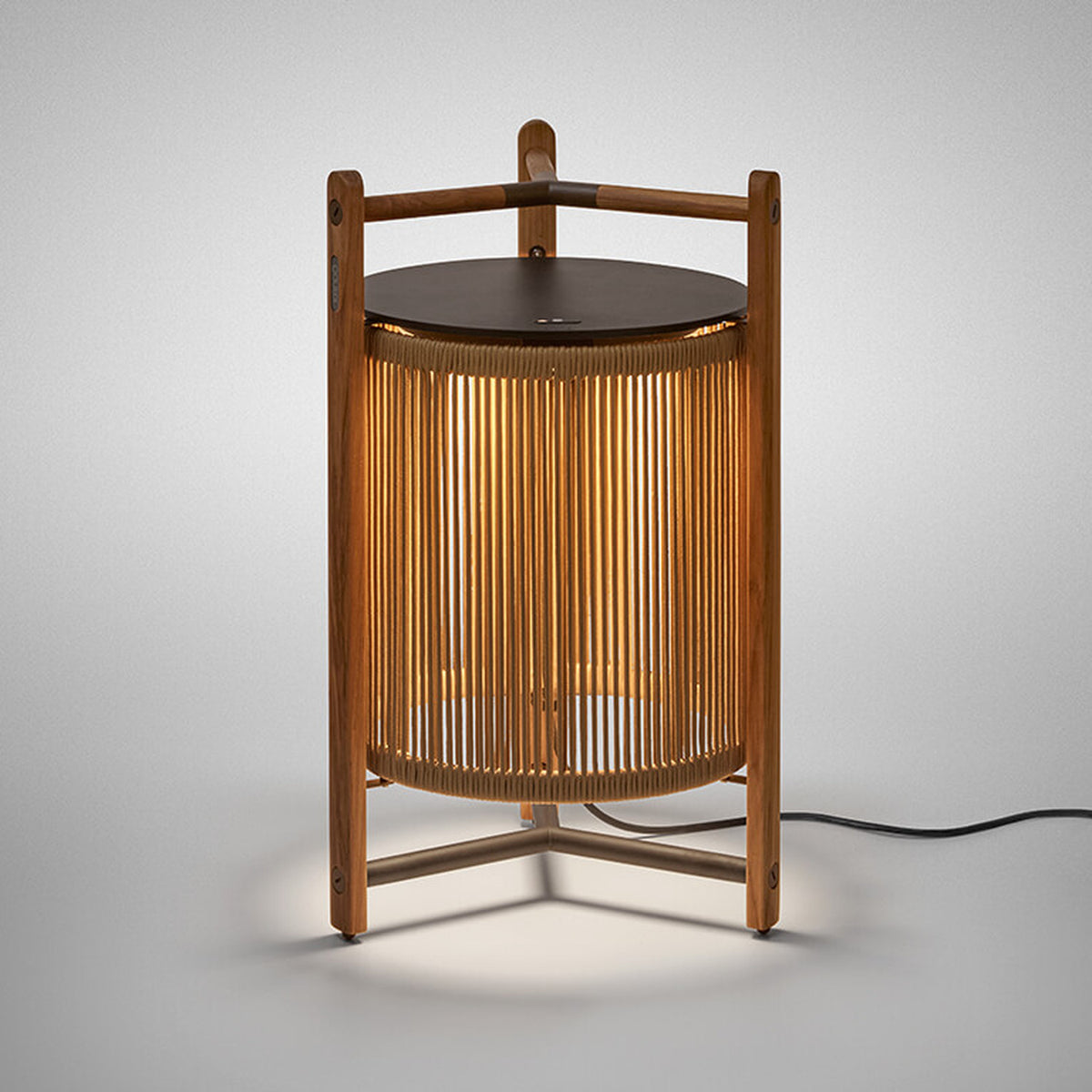 Kando P/76 outdoor floor lamp, lantern-style with sand nautical cord cylinder, teak wood tripod frame, dark round top and handle, illuminated, on a plain light grey background.