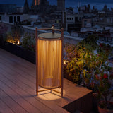 Kando P/111/R rechargeable outdoor floor lamp with sand nautical cord cylinder and teak wood frame, glowing on a rooftop terrace at night.