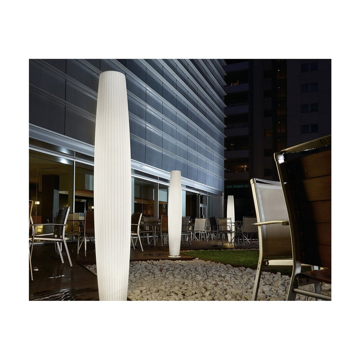 Maxi P/180 cylinder LED outdoor floor lamp in white polyethylene with ribbed texture, illuminated at night on a terrace beside modern buildings and patio seating.