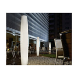 Maxi P/180 cylinder LED outdoor floor lamp in white polyethylene with ribbed texture, illuminated at night on a terrace beside modern buildings and patio seating.