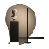 Solid F 180 cylinder LED floor lamp with a slim black upright column and round flat base, shown beside a classical bust on a dark plinth against a neutral backdrop.