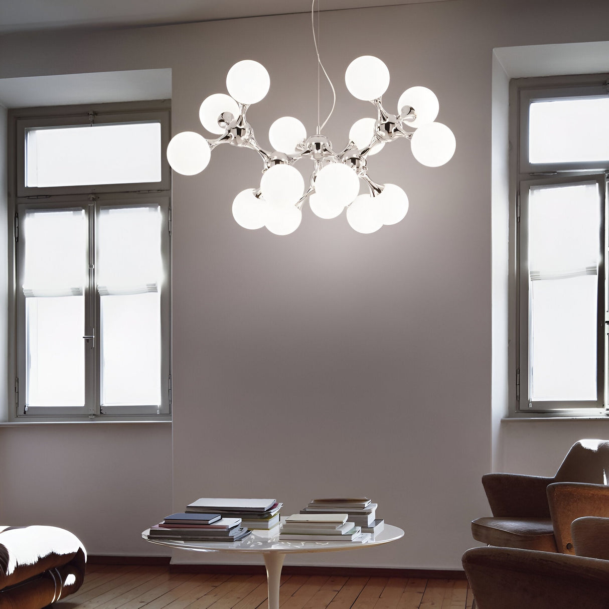 In a modern room with large windows, the Emblaze 15 Light Suspension - White graces the space, its frosted glass shades lending an exquisite touch. Beneath it, a round table with stacks of books is positioned between two armchairs on the wooden floor, perfectly embodying contemporary style and elegance.