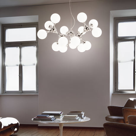 In a modern room with large windows, the Emblaze 15 Light Suspension - White graces the space, its frosted glass shades lending an exquisite touch. Beneath it, a round table with stacks of books is positioned between two armchairs on the wooden floor, perfectly embodying contemporary style and elegance.