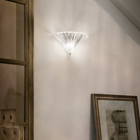 A Spring Large Glass Wall Light in clear glass emits soft light in a room characterized by a classic, elegant design. A framed architectural drawing leans against the wall, and a tufted chair is partially visible on the left. This warm setting achieves a sophisticated blend of timeless charm and artisanal flair.