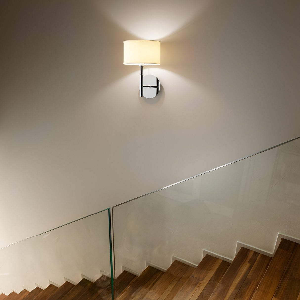 A modern staircase with wooden steps and a glass railing is highlighted by the elegant Noble Light Wall Light - Cream. Its soft glow illuminates the pristine white wall and sleek chromed metal frame, creating a warm and welcoming ambiance.