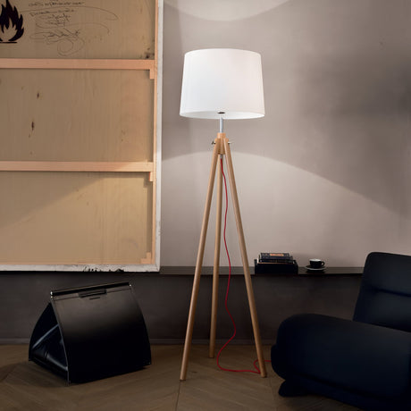 A modern living room embodies minimalist interiors, featuring the Blues Wood Floor Lamp with a tall wooden tripod base and a white shade accented by a red cord. A black chair sits prominently in the foreground, while a small table holds a cup and book. On the wall, part of a framed canvas with scribbles adds an artistic touch.