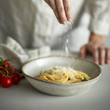 Sandrine Stoneware Deep Plate in light grey speckled glaze with pasta and grated cheese, shown as someone sprinkles seasoning, with cherry tomatoes beside it on a kitchen counter.