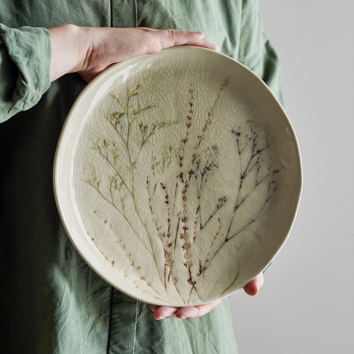 Bea Crackle Stoneware Dinner Plate in cream, round with gently irregular rim, glossy crackle glaze and floral grass motifs, held in two hands against a green garment.
