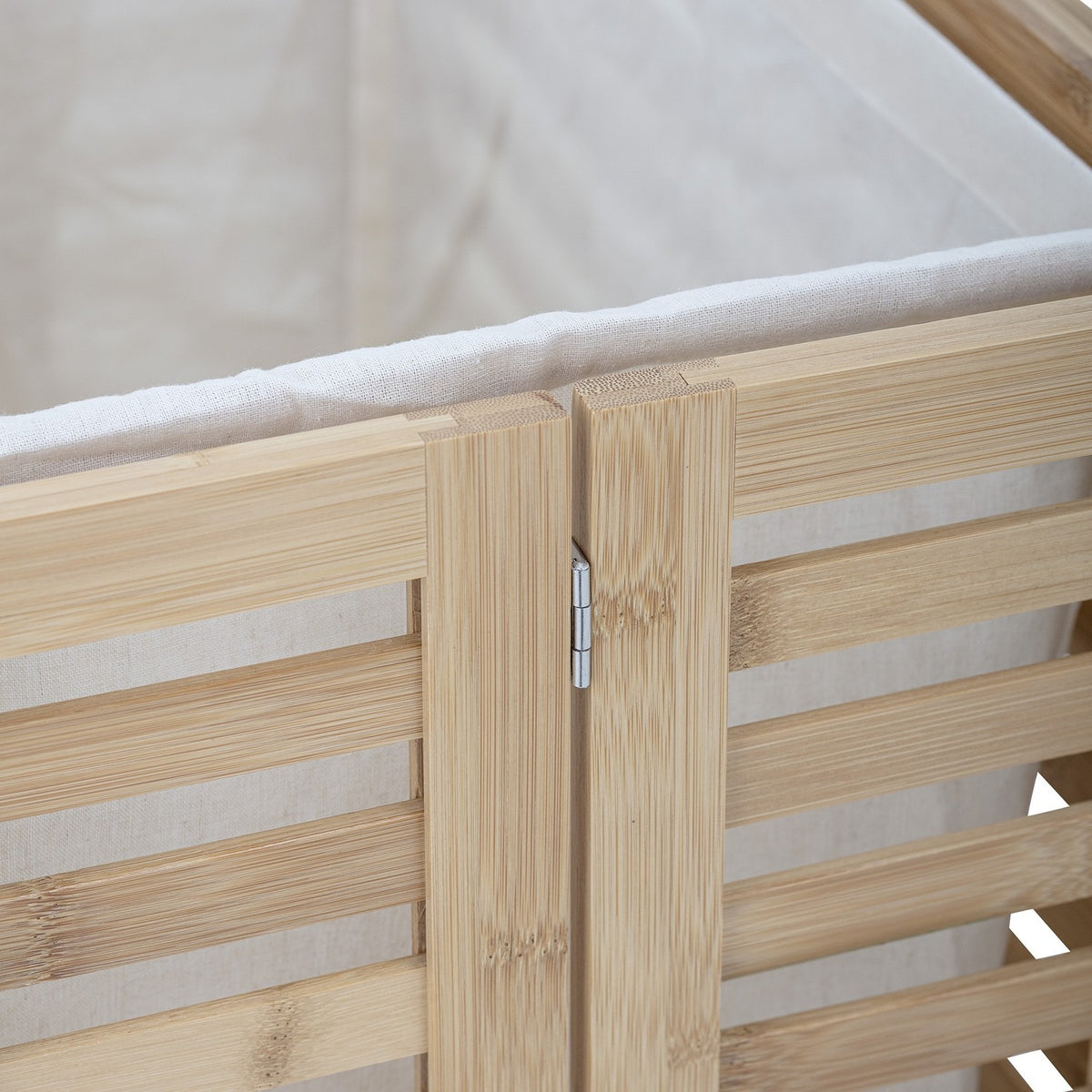 Close-up of the Aden Bamboo Laundry Basket's hinged lid corner, showing natural slatted bamboo panels, a metal hinge, and the cream cotton liner folded over the rim.