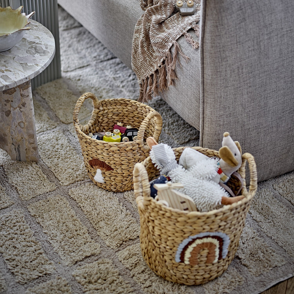 Agnes Water Hyacinth Baskets Set of 2 in beige, round woven storage baskets with handles, one with a stitched mushroom and one with a rainbow motif, filled with children's toys on a living room rug.