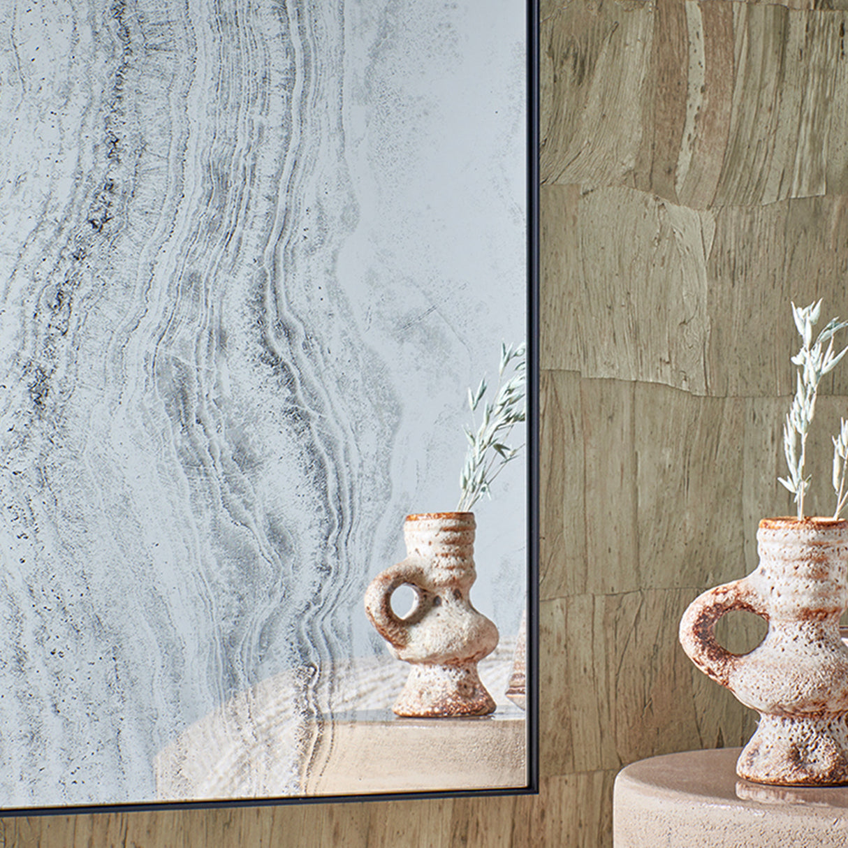 Close-up of the mirror at an angle beside ceramic jugs, the grey stone print set against a textured stone wall.