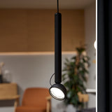 Berry S Aluminium LED Pendant Light in matte black, slim tube with an angled domed head, shown switched on above a dining area with a plant and armchair in the background.