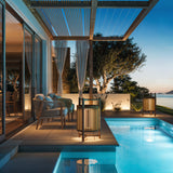 Kando P/76 Ribbon LED floor lamp with cream pleated cylinder shade on a teak three-legged frame, shown on a poolside patio beside lounge seating at dusk.