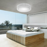 White round Urban PF/60 Ribbon LED flush ceiling light with pleated drum shade, mounted on concrete above a bedroom with a wooden bed and glass wall.