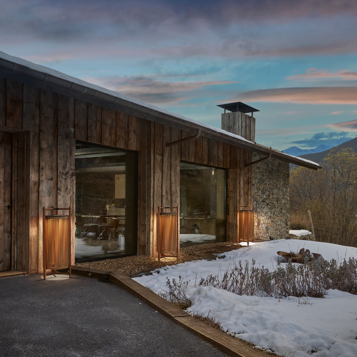 Kando P/111 wood outdoor floor lamps with cylindrical nautical cord shades in teak frames, illuminated beside a rustic wood-clad house at dusk with snow-covered ground.