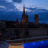 Nans Bag/38/R rechargeable outdoor floor lamp with woven beige basket shade and carry handles, shown lit on a rooftop terrace at dusk beside a cathedral skyline.