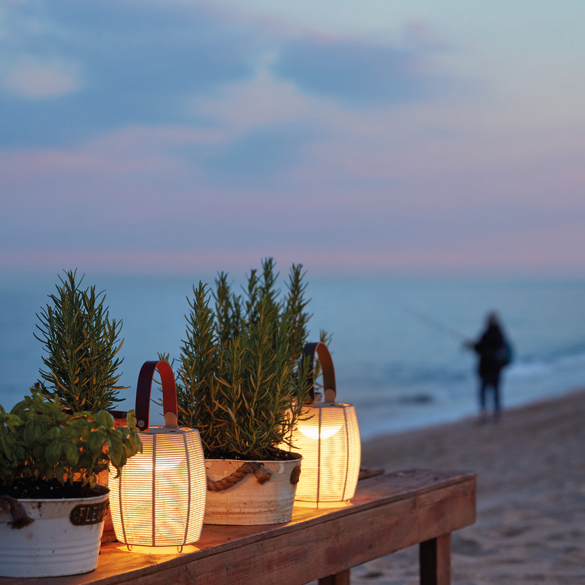 Tanit M/29/R rechargeable table lamps with woven brown shades and carry handles, glowing beside potted herbs on an outdoor table at dusk by the beach.
