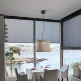 Syra 45 Aluminium LED Pendant Light with a woven ivory drum shade narrowing at the waist, hung above a dining table in a seaside living room with roller blinds and sea views.