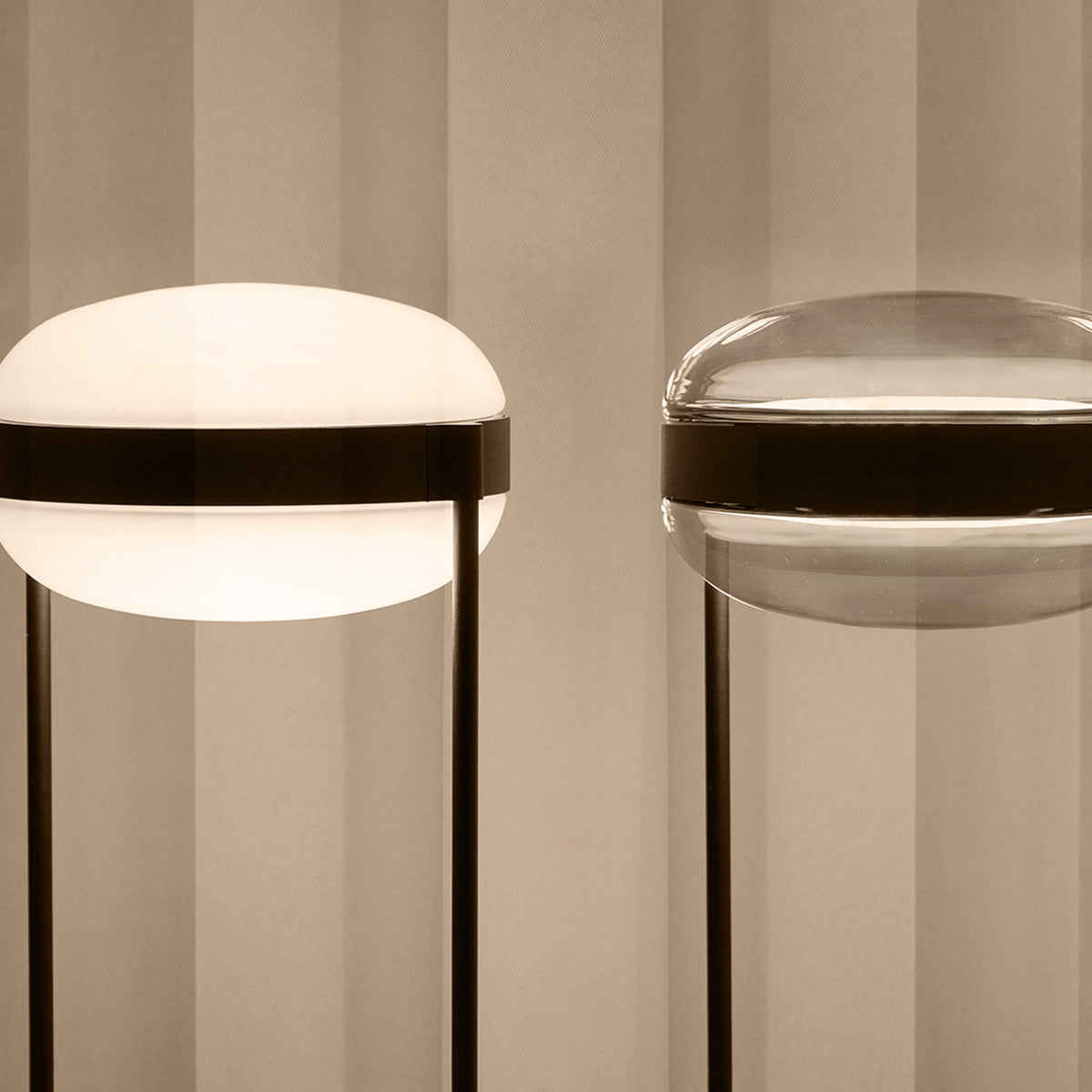 Beaded T Glass LED table lamps with flattened spherical shades in opal white (lit) and smoke grey, each with a dark centre band and slim rod legs on a round base.
