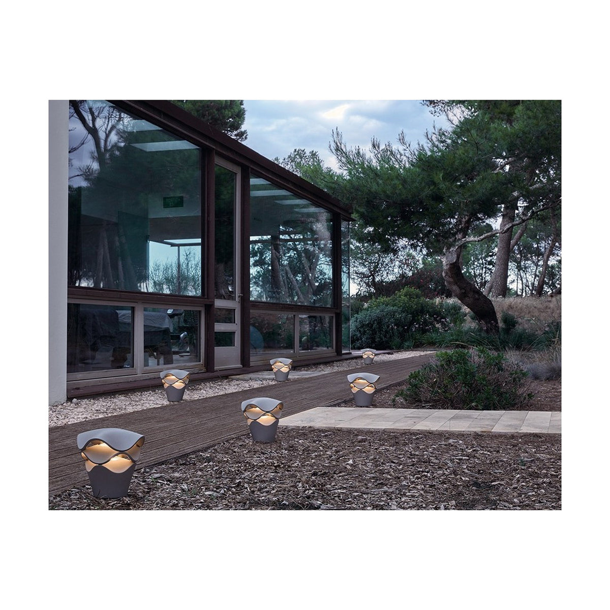 Cornet B/27 Cone LED bollard lights in Stone Grey with wavy tiered shades, glowing warm white along a garden path beside a glass-fronted house.
