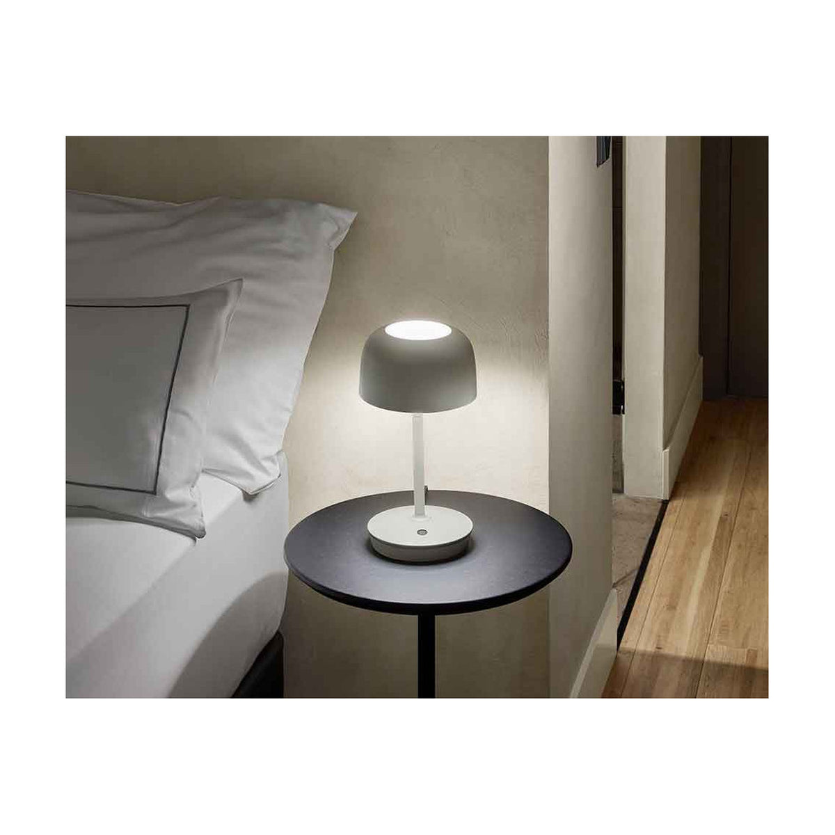 Bover Bol M/37 dome LED table lamp in grey with slim stem and round base, glowing warmly on a dark bedside table beside a bed.