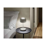 Bover Bol M/37 dome LED table lamp in grey with slim stem and round base, glowing warmly on a dark bedside table beside a bed.
