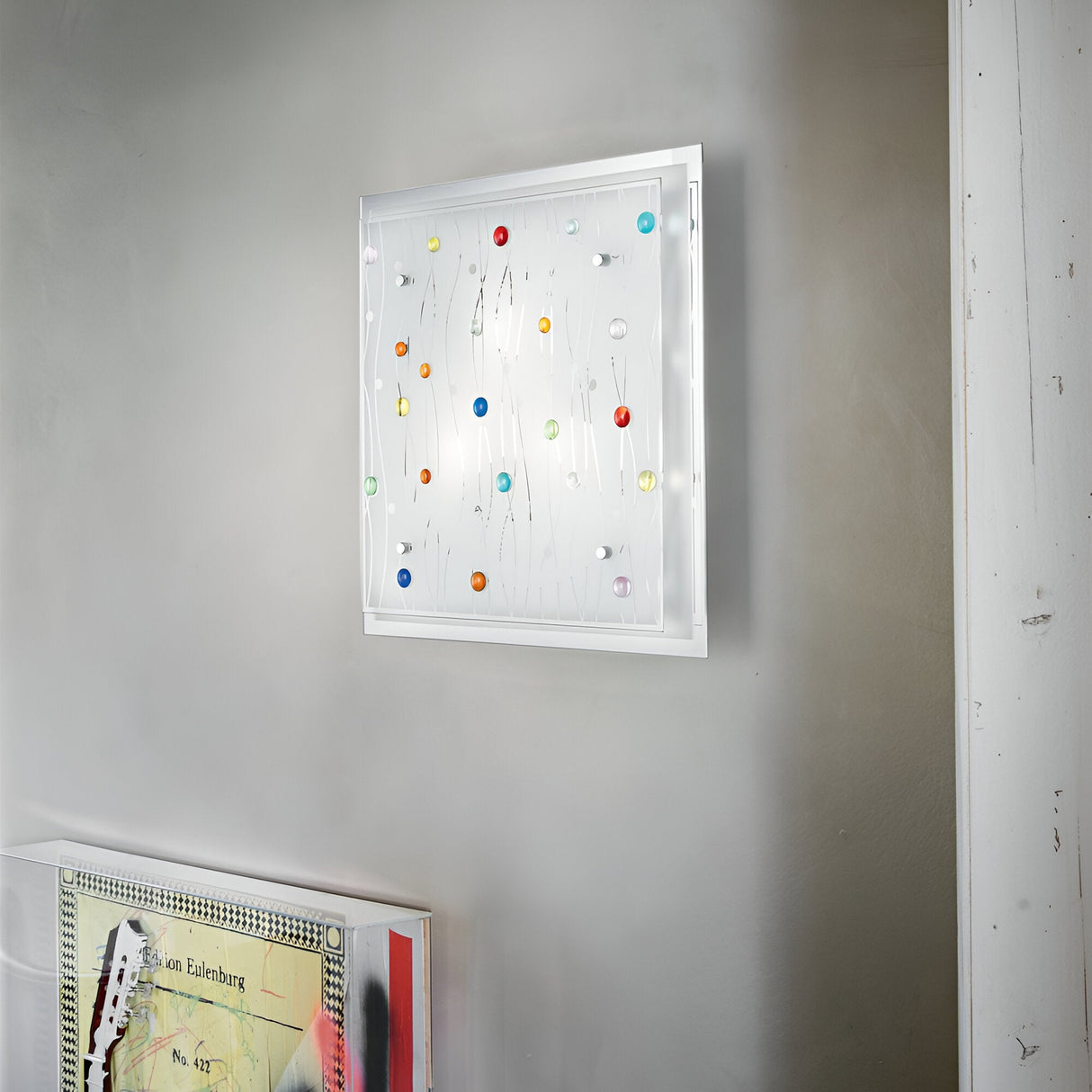 A contemporary art piece adorns the wall, showcasing a white background with vibrant, abstract shapes in shades of red, blue, yellow, and green. Dynamic lighting effects illuminate the artwork. Below it is a segment of another piece featuring geometric patterns in muted tones under the Horizon 3 Light Ceiling & Wall Light - Multicolour.