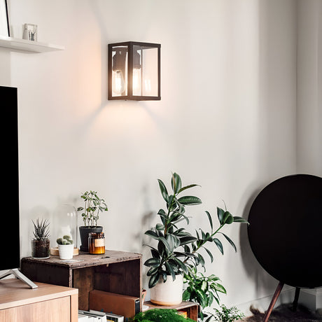 A cozy living room corner showcases modern lighting with Drape Wall Lights in black, surrounded by various potted plants. A round black object leans against the wall, and a TV is partially visible. The decor incorporates wooden and glass elements, creating a natural and inviting ambiance.