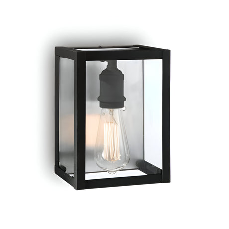 The Drape Wall Lights - Black is a contemporary lighting fixture with a sturdy black metal rectangular frame and a clear glass enclosure, housing an exposed vintage-style Edison bulb. Mounted on a white wall, it offers both style and timeless appeal.