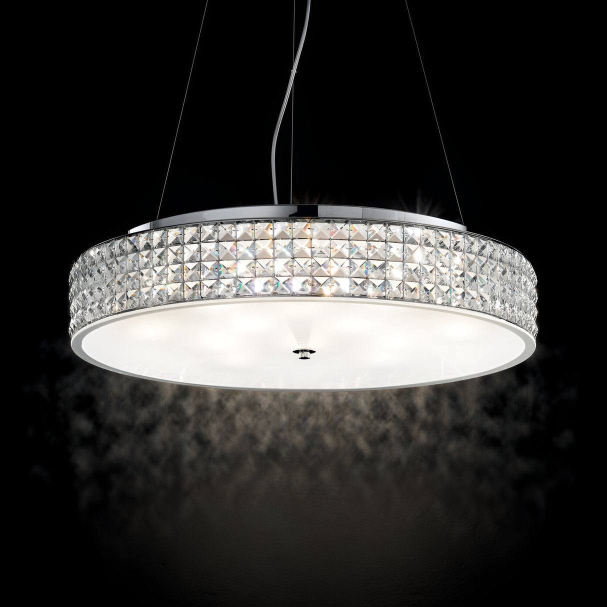 The Nebula 12 Light Crystal Suspension - Chrome features a circular metallic frame with crystal-like enhancements. This chrome chandelier hangs from adjustable suspension cables against a dark backdrop, providing a gentle, white illumination.