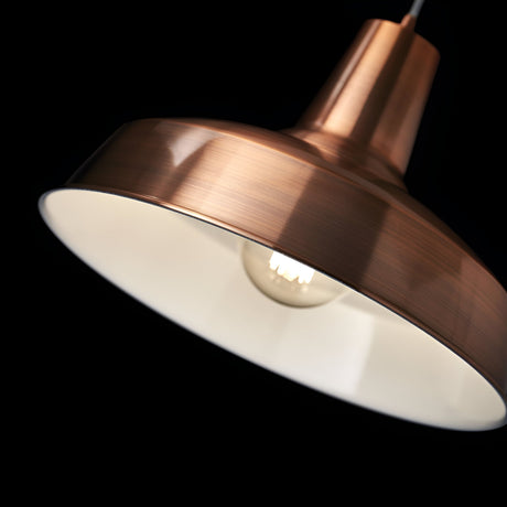 A close-up of the Blaze 1 Light Pendant in copper against a dark background. This modern industrial home decor piece is illuminated, highlighting the bulb inside and its dimmable lighting feature that casts a warm glow on the white interior of the shade.