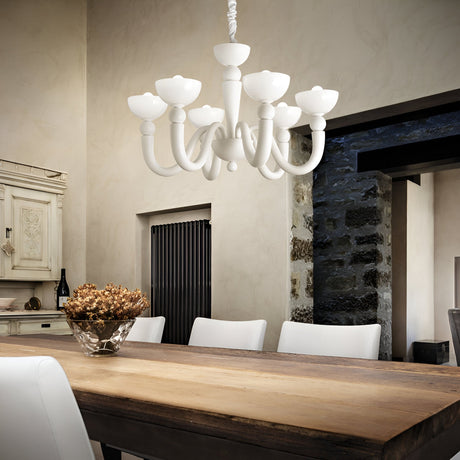 A rustic dining room showcases a wooden table surrounded by white chairs, illuminated by the elegant Swift Glass 8 Light Chandelier - White. A bowl of dried flowers adorns the table, while cream-colored walls and a blend of stone and wood elements enhance the inviting decor.