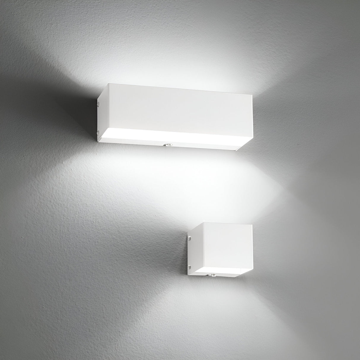 Two Vividara Wall Lights in white, embodying minimalist lighting and modern design, are mounted on a gray wall. They cast soft white light both upwards and downwards. The larger fixture positioned above the smaller one creates intersecting beams that form triangular patterns on the wall.